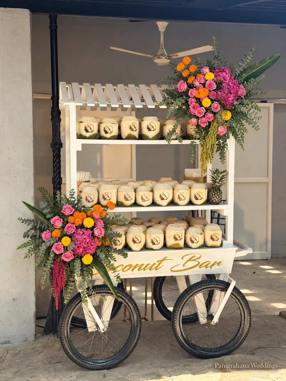 A white coconut bar cart on bicycle wheels, its shelves of tender coconuts dressed with pink and orange flower bunches, at a haldi in Bangalore