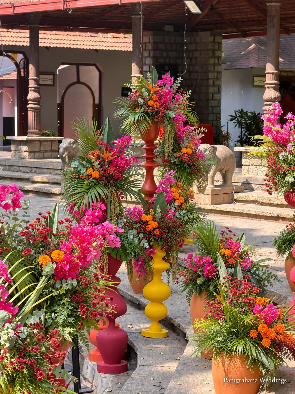 Courtyard steps at The Tamarind Tree lined with tall arrangements of pink bougainvillea, heliconia and palms in red and yellow painted urns, with stone elephants at the entrance