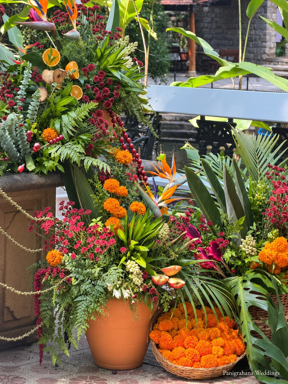 Tall tropical arrangement with dried orange slices, bird of paradise, burgundy chrysanthemums, ferns, monstera and bead strands, over a terracotta pot and a basket of marigold heads