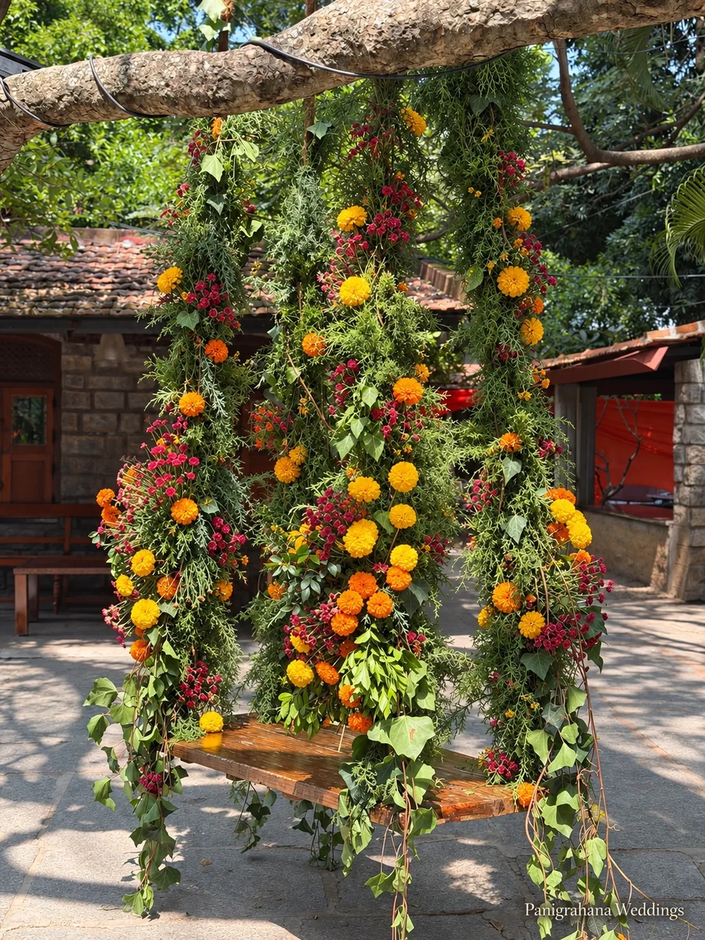 A wooden swing hung from a tree branch on ropes wrapped in foliage, marigolds and red flowers, set up in the courtyard for a haldi at The Tamarind Tree