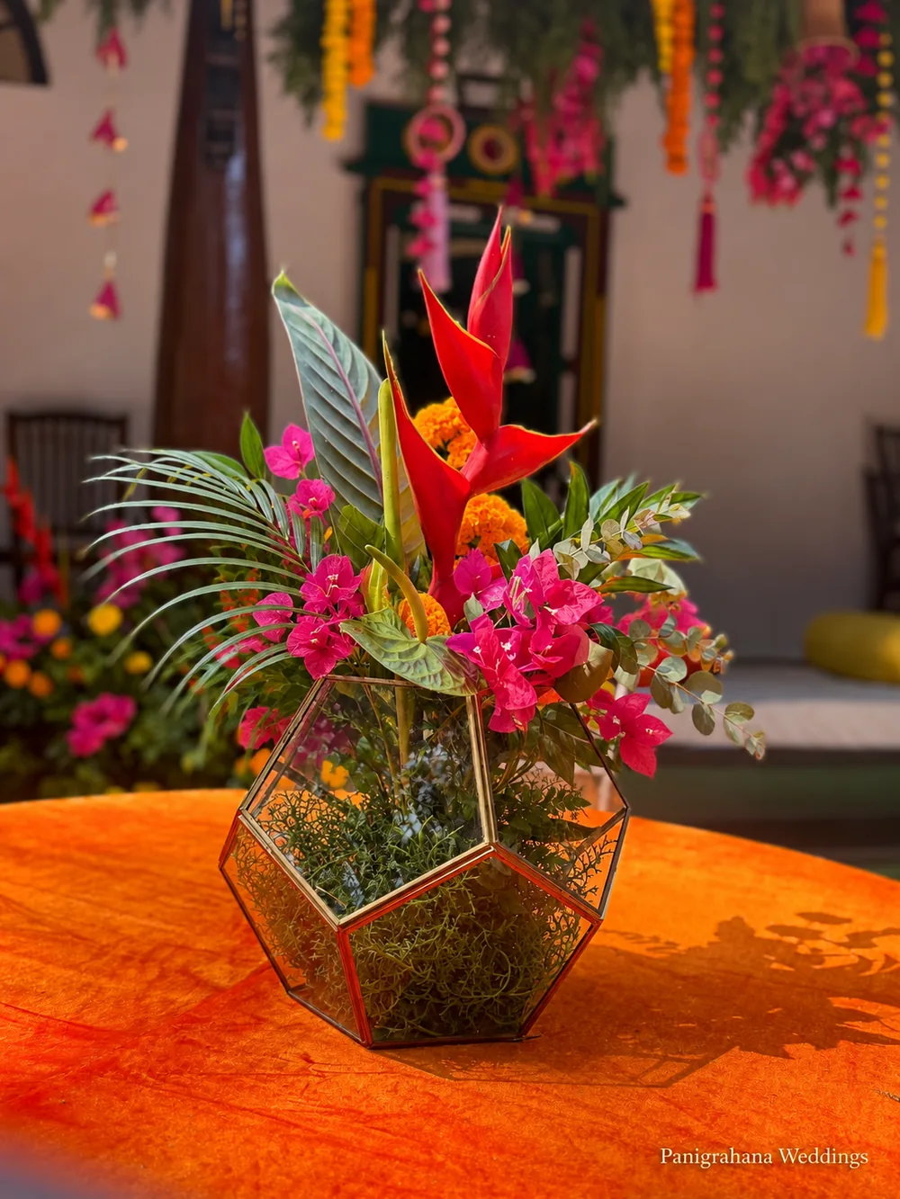 Haldi table centrepiece: a brass-framed geometric glass terrarium holding red heliconia, pink bougainvillea and palm on an orange velvet table