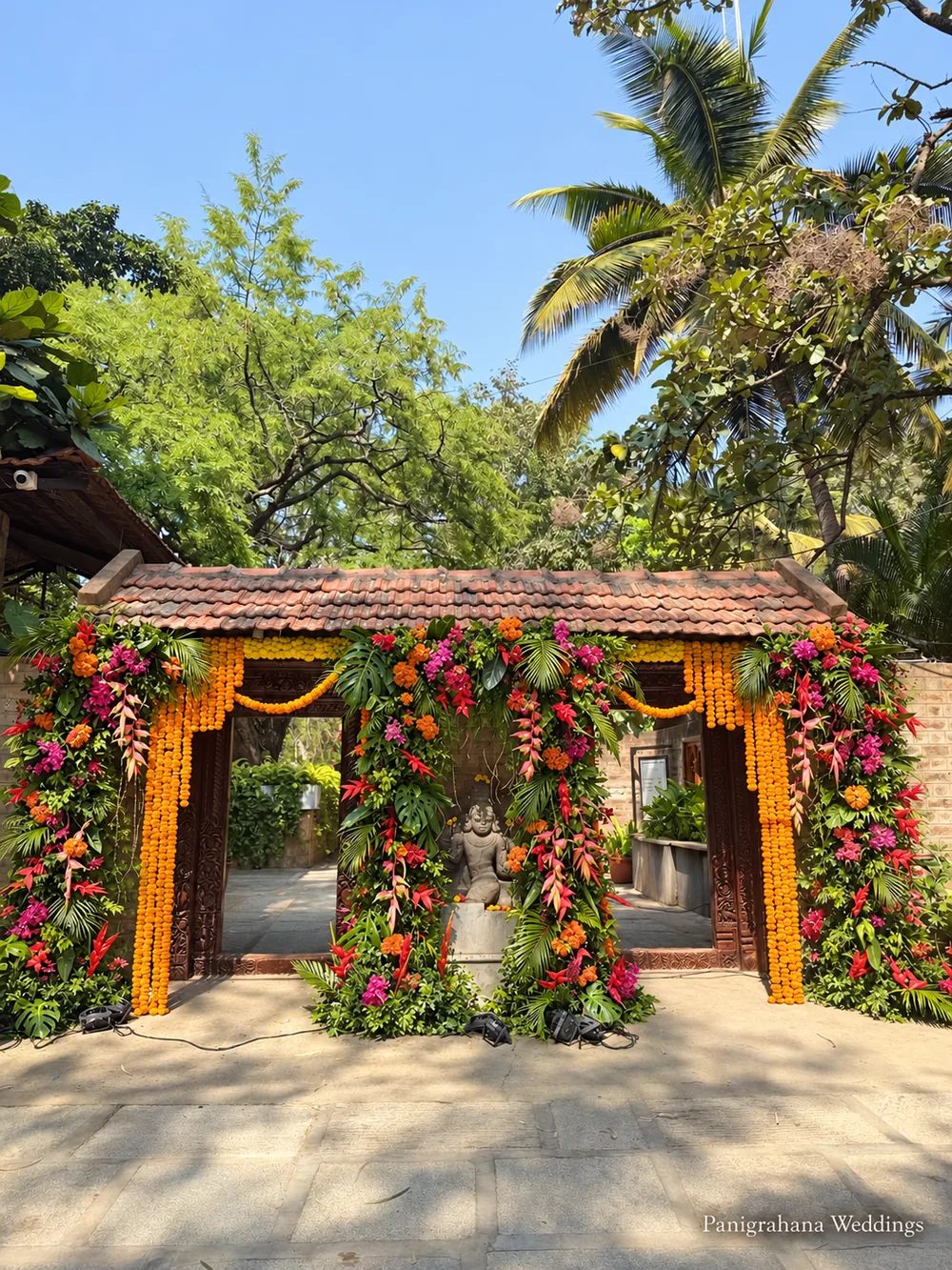 Tiled-roof entrance gate at The Tamarind Tree, Bangalore, its pillars wrapped in marigold strings and tropical foliage for a daytime haldi designed by Panigrahana