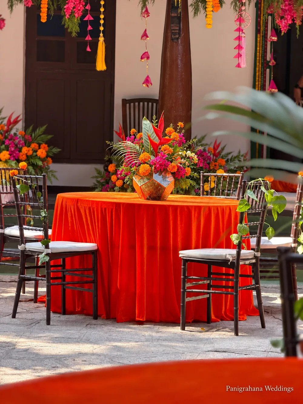 Haldi lunch table with an orange velvet cloth, dark Chiavari chairs with white cushions and a tropical centrepiece, under hanging marigold and pink garlands