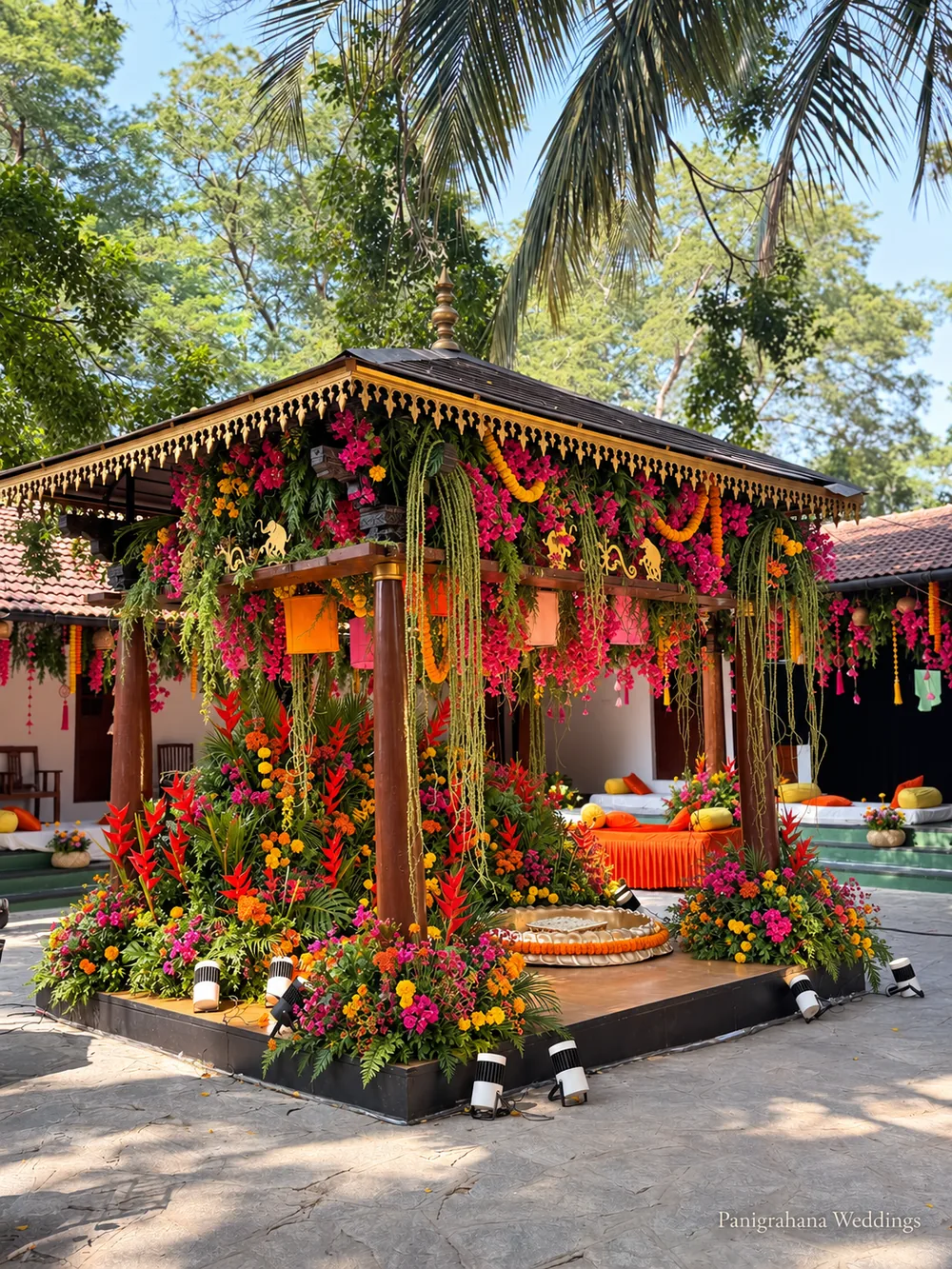Wide view of the haldi pavilion at The Tamarind Tree with hanging foliage and flowers on every side, low seating around it and tall palms behind