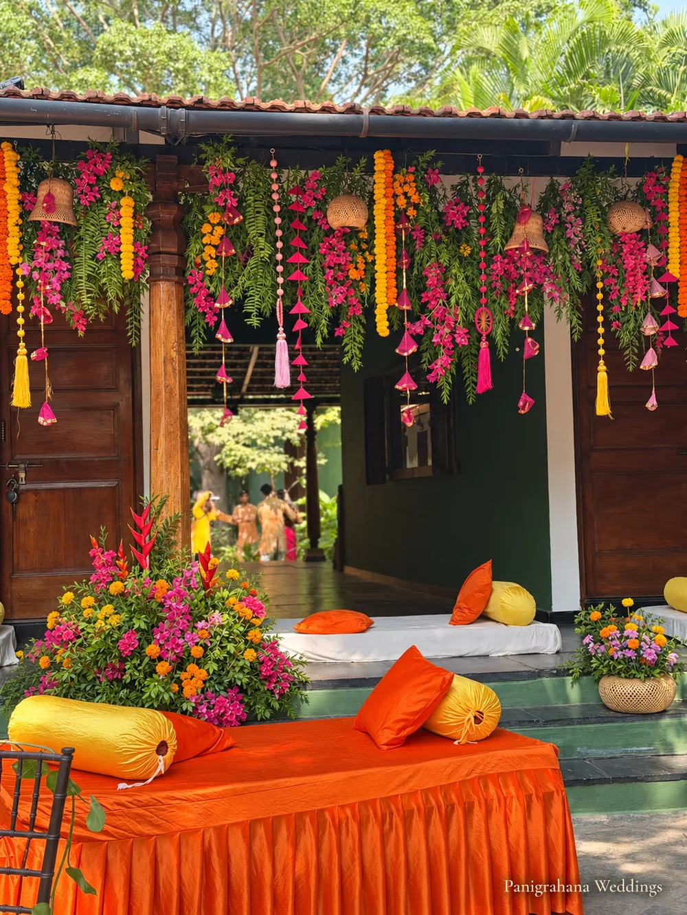 Low verandah seating for haldi guests with orange covers, yellow bolsters and orange cushions under hanging marigold and bougainvillea garlands