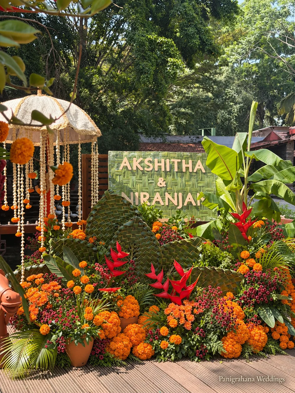 Haldi welcome corner with a woven coconut-frond name sign, a cream chhatri hung with marigold and jasmine strings, red heliconia, banana leaves and mounds of marigold in terracotta pots
