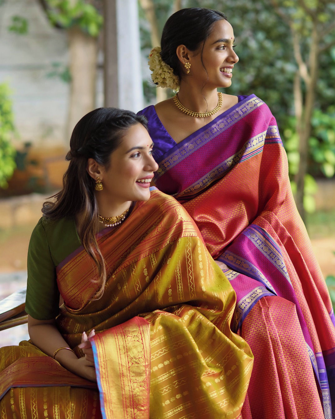Two women in silk sarees, one rust and gold and one purple with a red border, seated outdoors