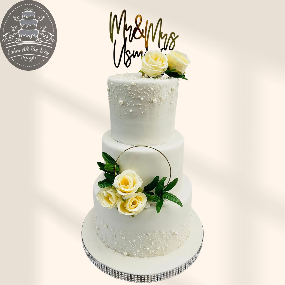 Three-tier white fondant wedding cake with pearl detailing, cream roses, a gold hoop and a Mr & Mrs topper