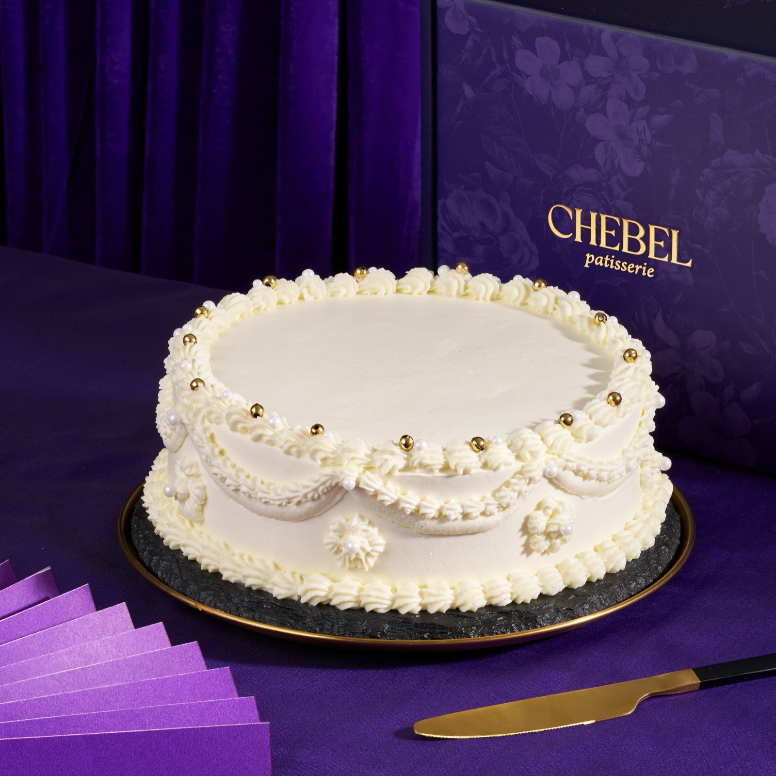 Round cake in white chantilly cream with piped swags, shell borders and gold and white pearls, beside a Chebel box