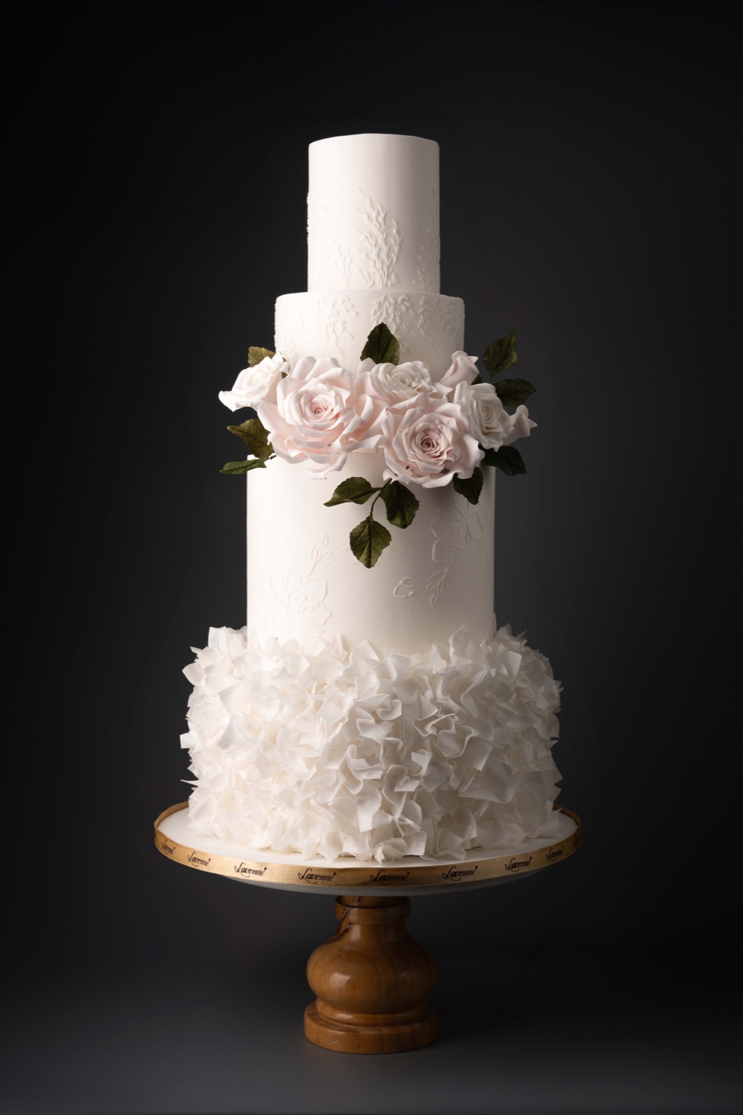 Four-tier white cake with a band of blush roses and leaves above a ruffled bottom tier, on a wooden stand