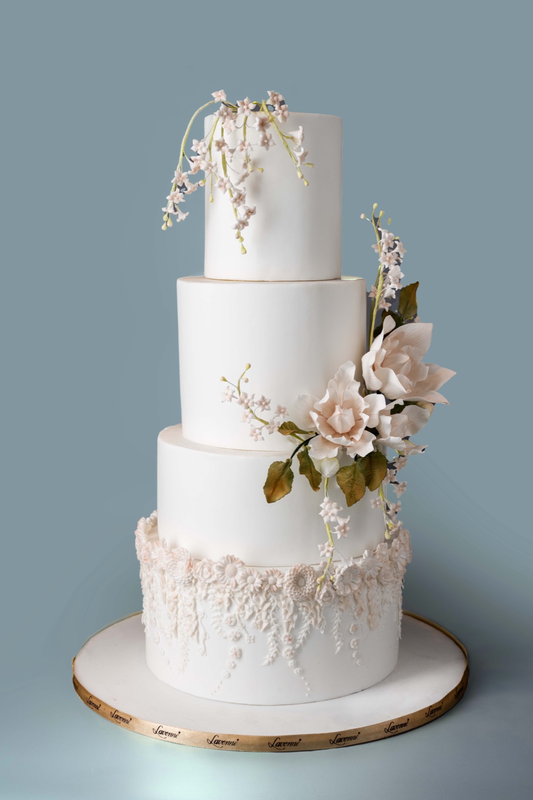 Four-tier white cake with pale pink flowers and leaves trailing down one side and lace-like piping at the base