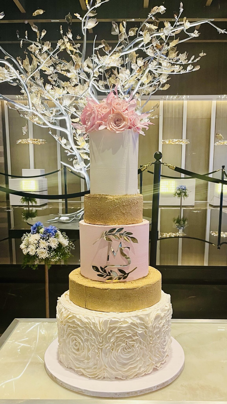 Five-tier cake with a ruffled white base, gold tiers, a pink monogram tier and pink roses on top