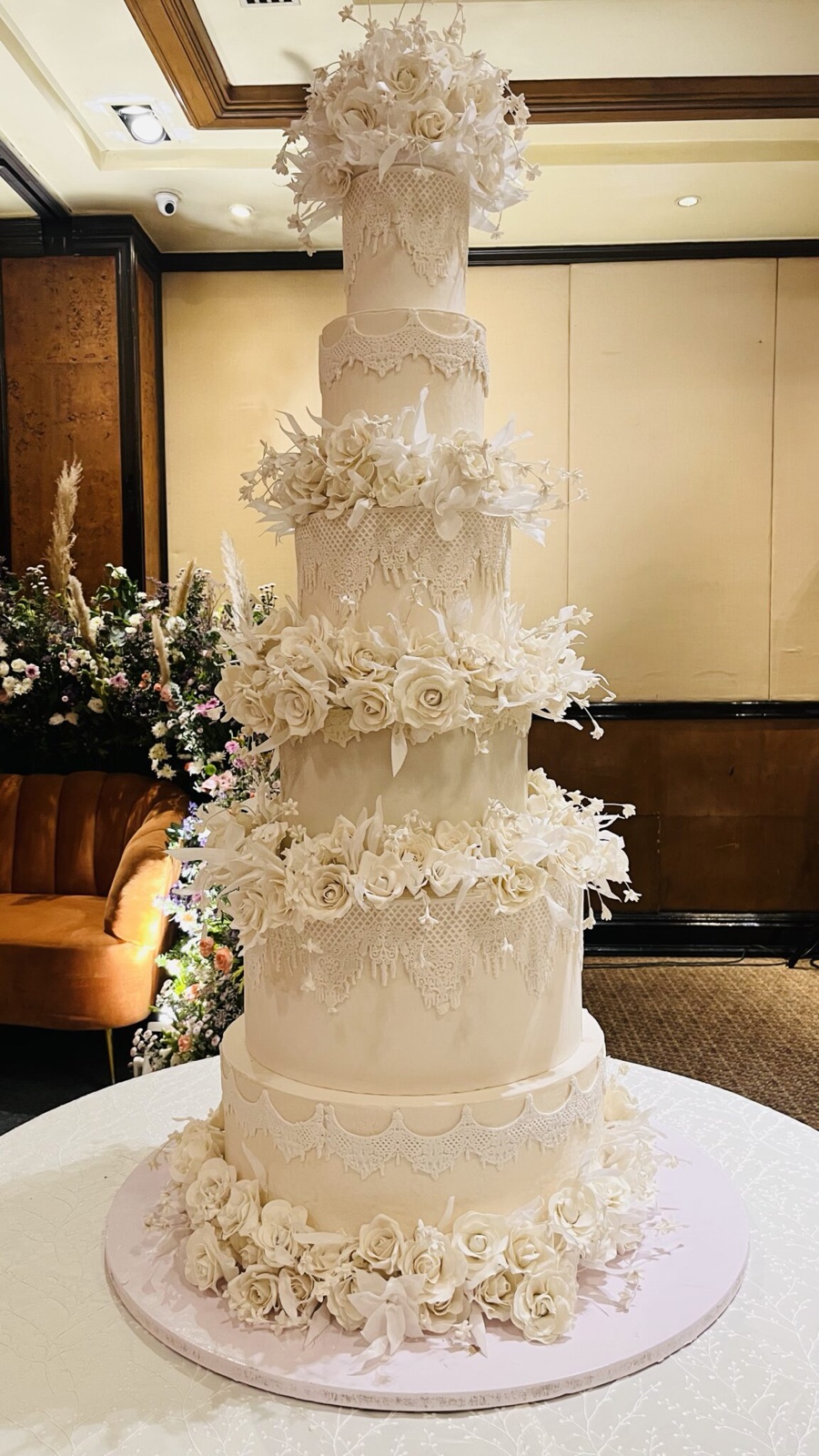 Tall all-white wedding cake with lace patterns and clusters of white roses between the tiers