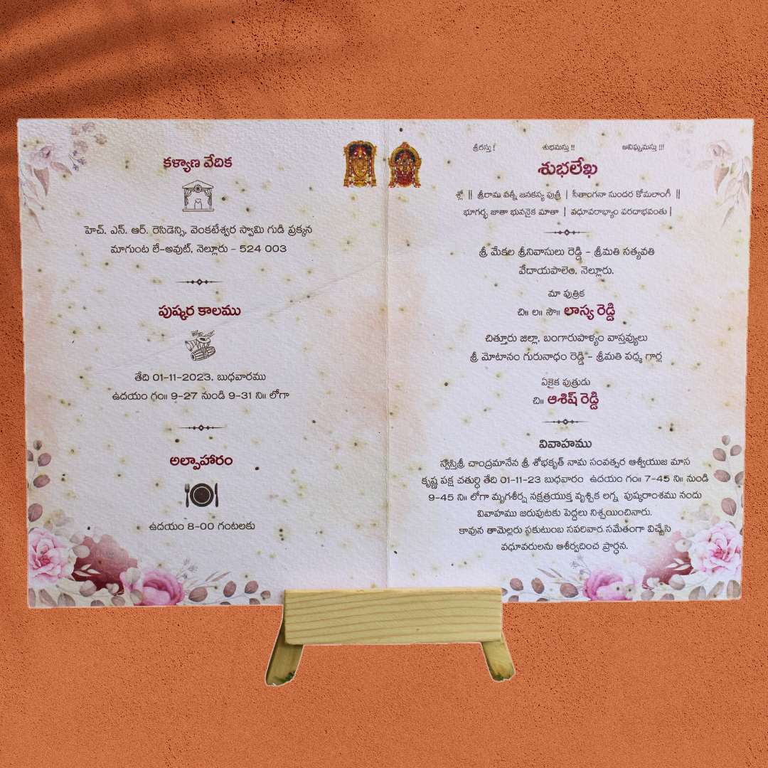 Seed-paper wedding card printed in Telugu with pink floral corners and seeds visible in the paper, on a small wooden stand
