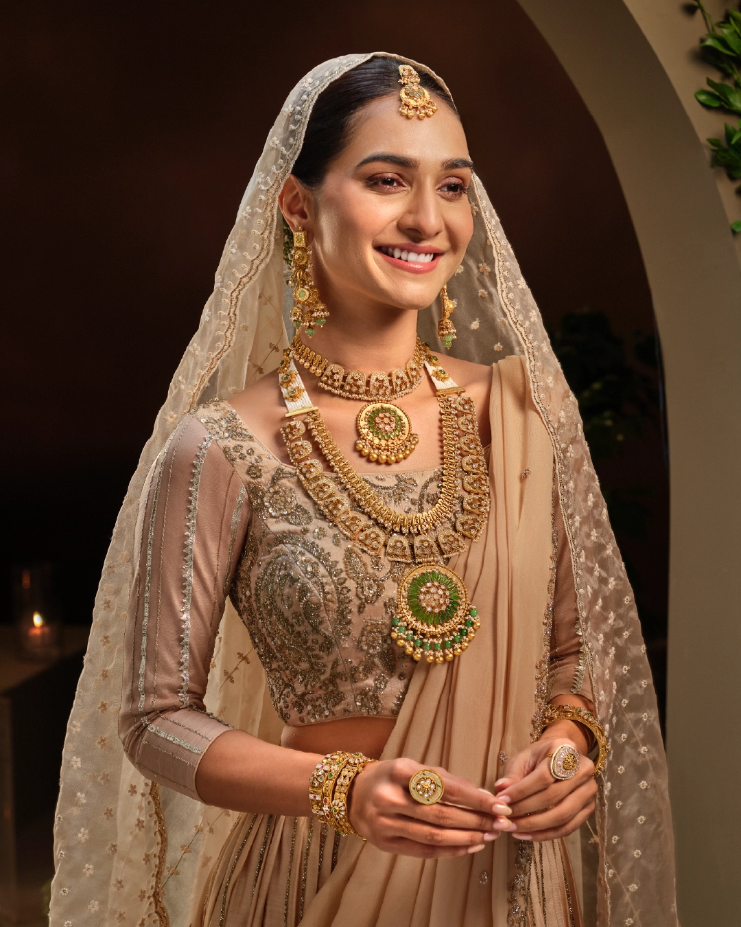 Bride in a beige lehenga wearing an antique gold choker, a long haram with green-stone pendants and a maang tikka