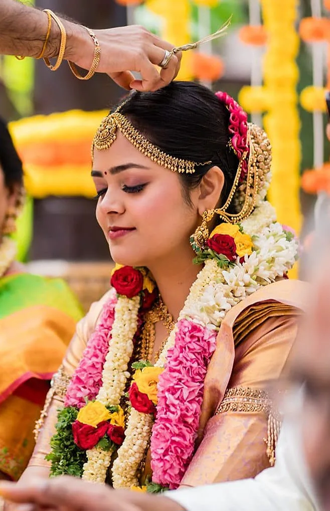 South Indian bride in a peach and gold silk saree with garlands, a gold headpiece and roses and jasmine in her hair during a ritual