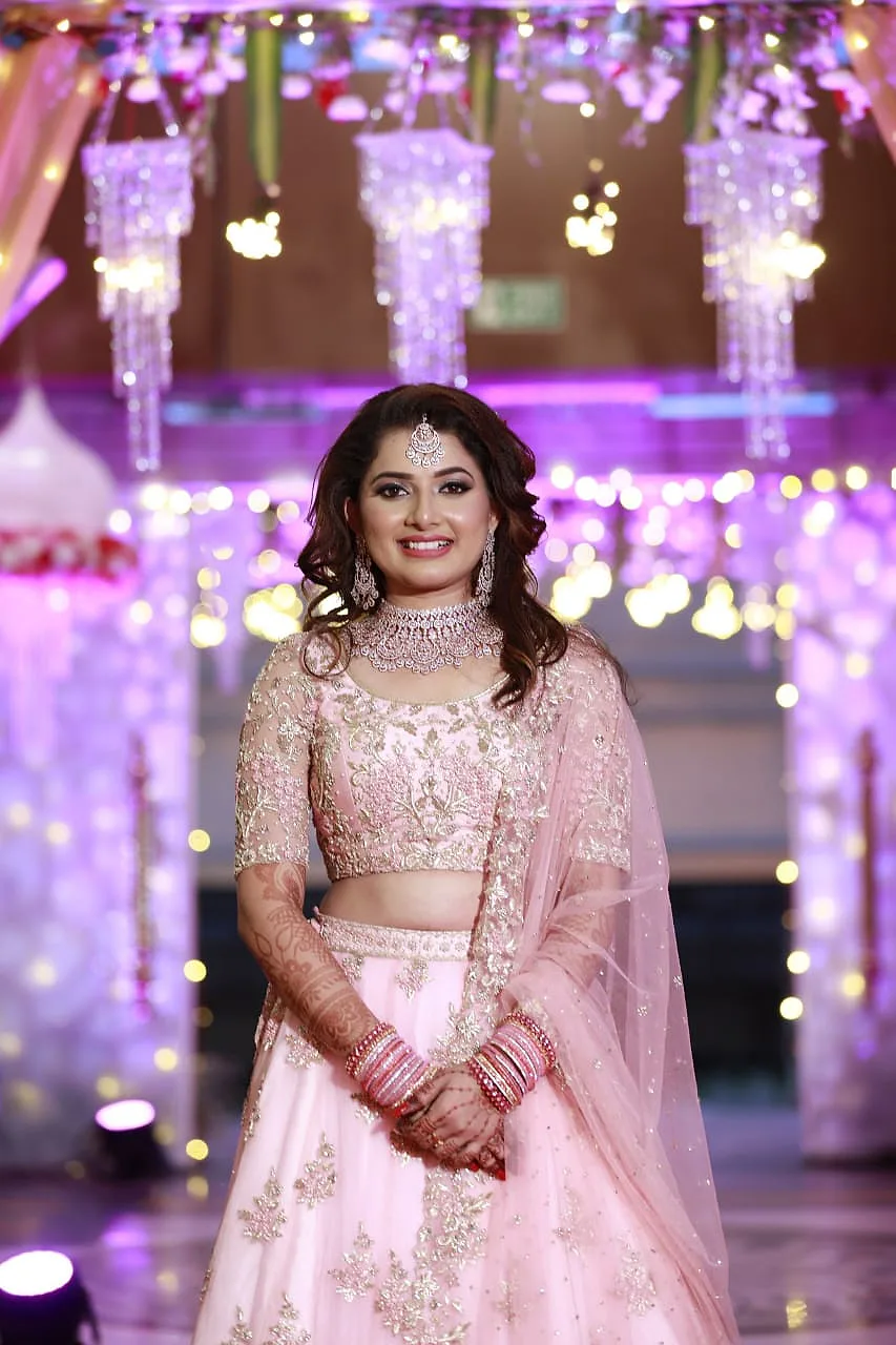 Bride in soft glam makeup and open curls, wearing a pale pink embroidered lehenga on a reception stage