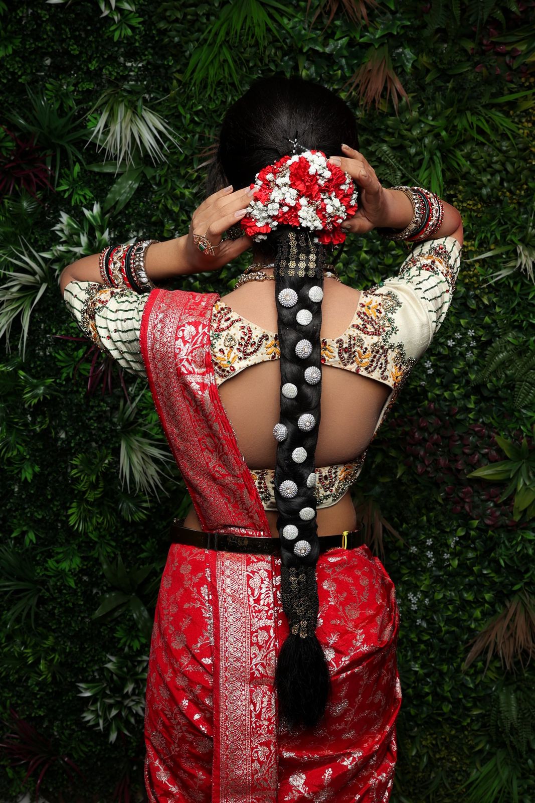 Back view of a bride's red and white flower bun above a long braid studded with silver ornaments, with a red silk saree