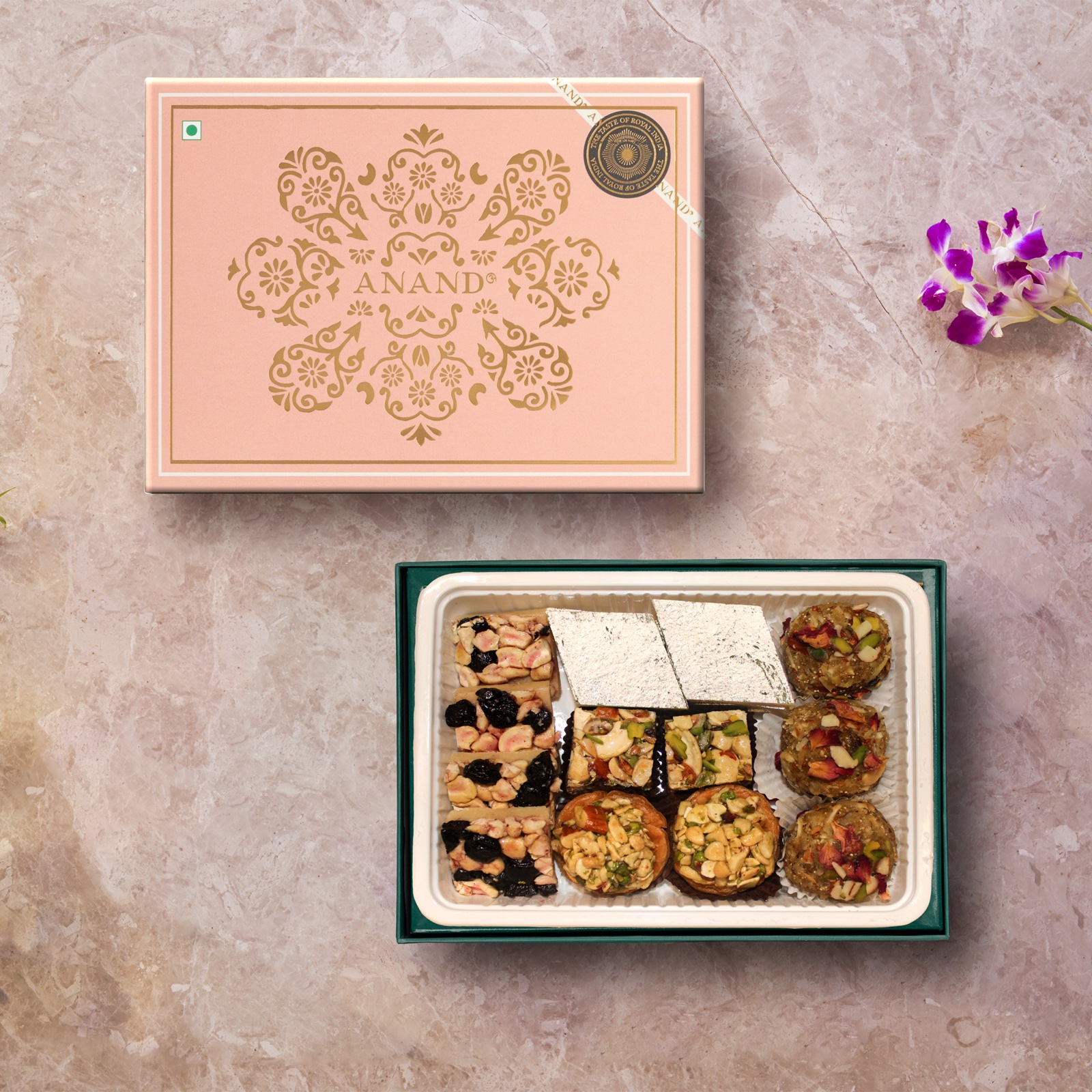 Open box of assorted sweets, including kaju katli and dry-fruit sweets, beside a pink Anand gift box