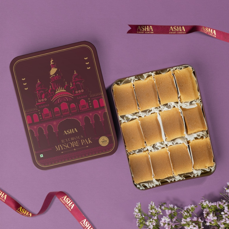 Open tin of Mysore Pak beside its maroon palace-print lid and Asha Sweet Center ribbon