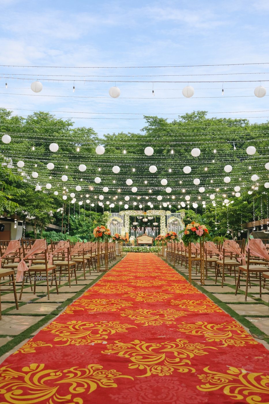 Lawn at Aarambha set with a patterned red aisle runner, gold chairs down both sides and white paper lanterns strung overhead against the sky