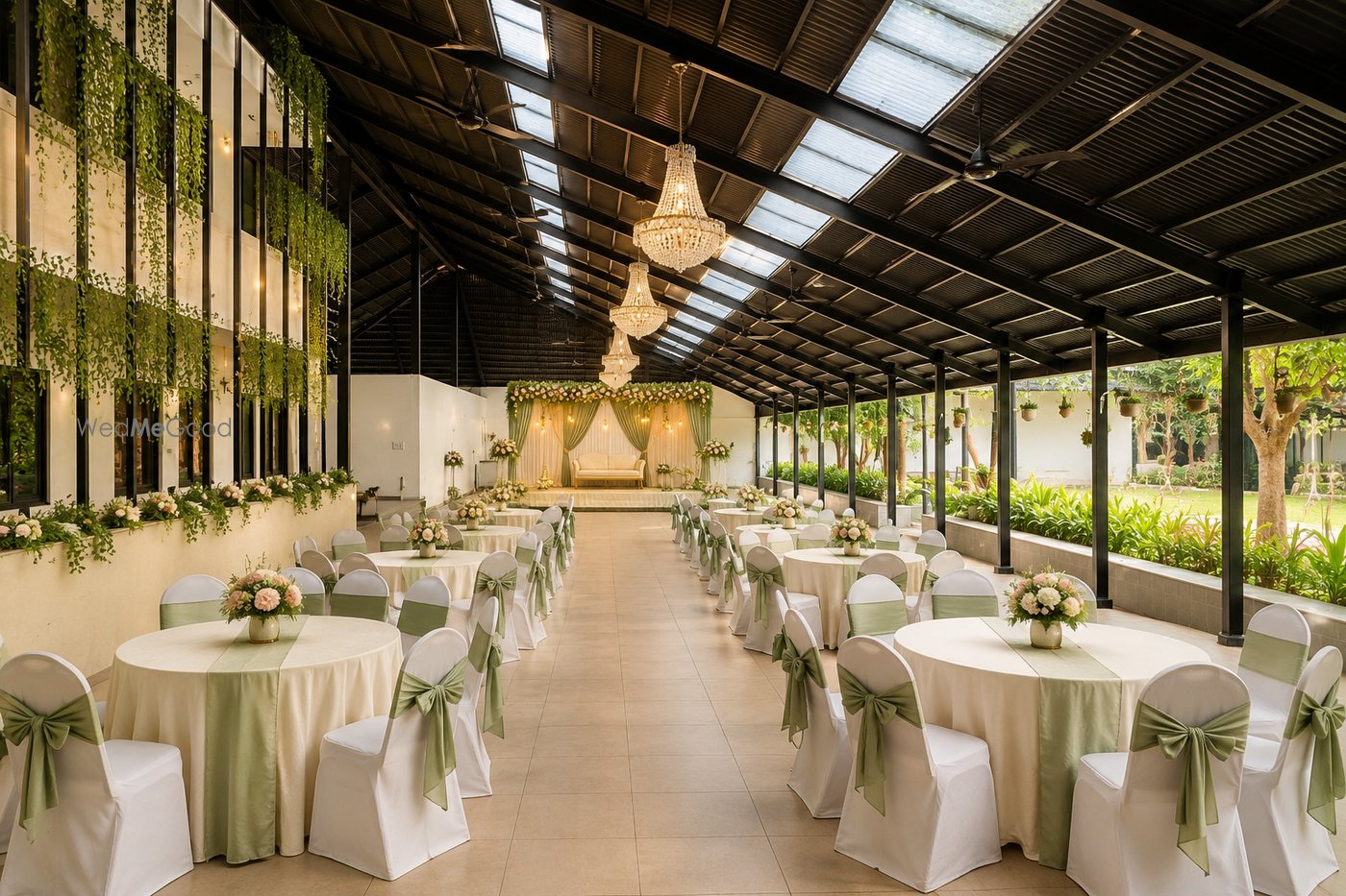 Banquet Hall 1 at Aarambha — a long room under a pitched steel roof with a vertical garden wall on the left, round tables in sage green and a chandelier down the spine