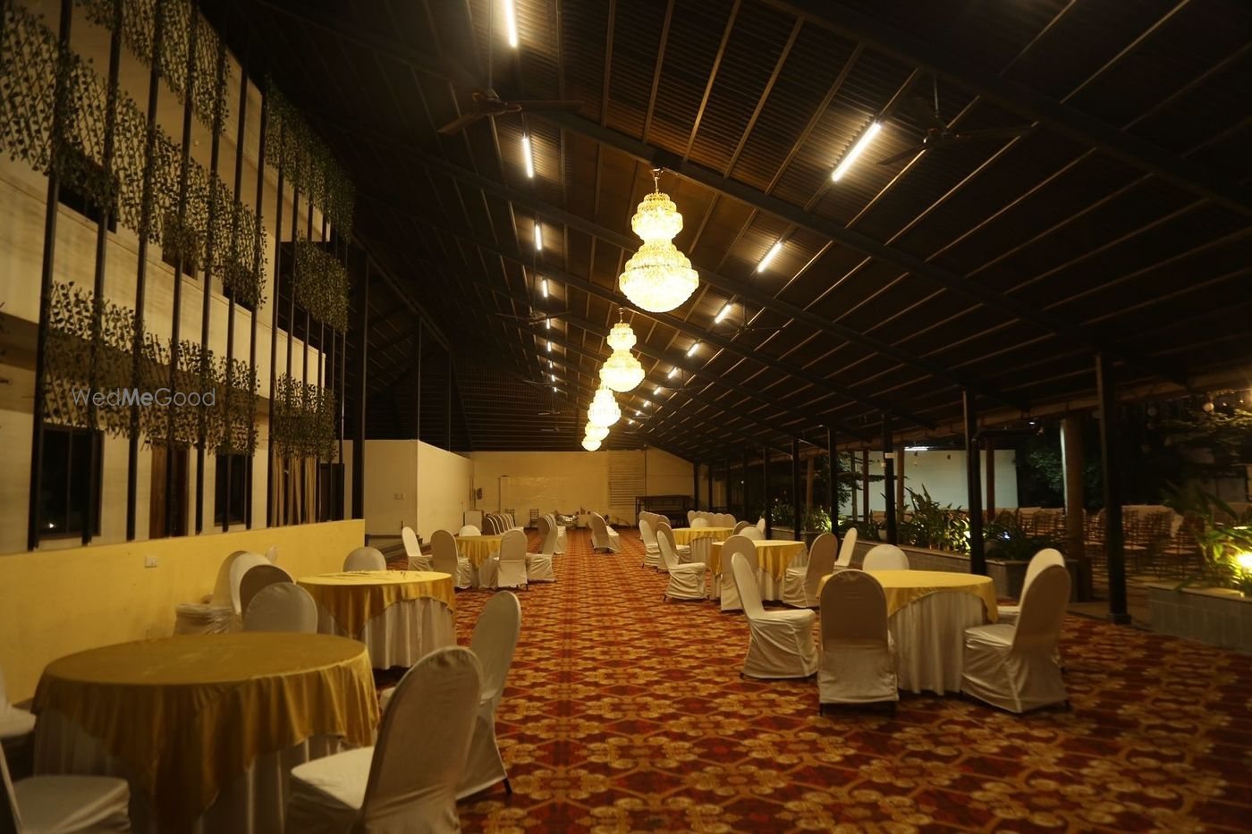 Dining hall at Aarambha under an exposed steel roof with warm lantern lighting, round tables in gold and a green wall along the far side