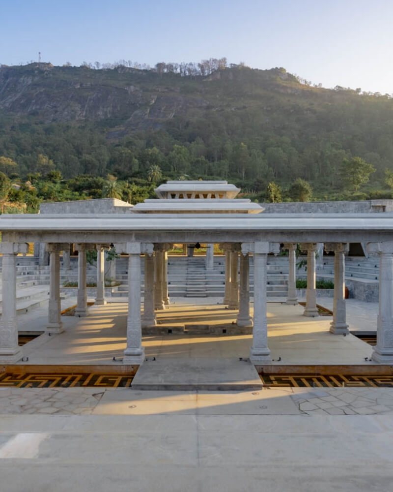 The Mantapa at Amita Rasa, undressed in morning light — a granite colonnade with a tiered pyramidal stone roof over the central altar, inlaid floor bands, the stepped seating of the Kalyani rising behind it and the wooded Nandi ridge above