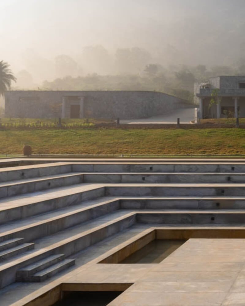The Kalyani amphitheatre at Amita Rasa empty in early-morning hill mist — wide stone steps descending on three sides to a sunken square floor, with the estate's stone boundary wall, a lawn and a villa beyond, all softened by fog
