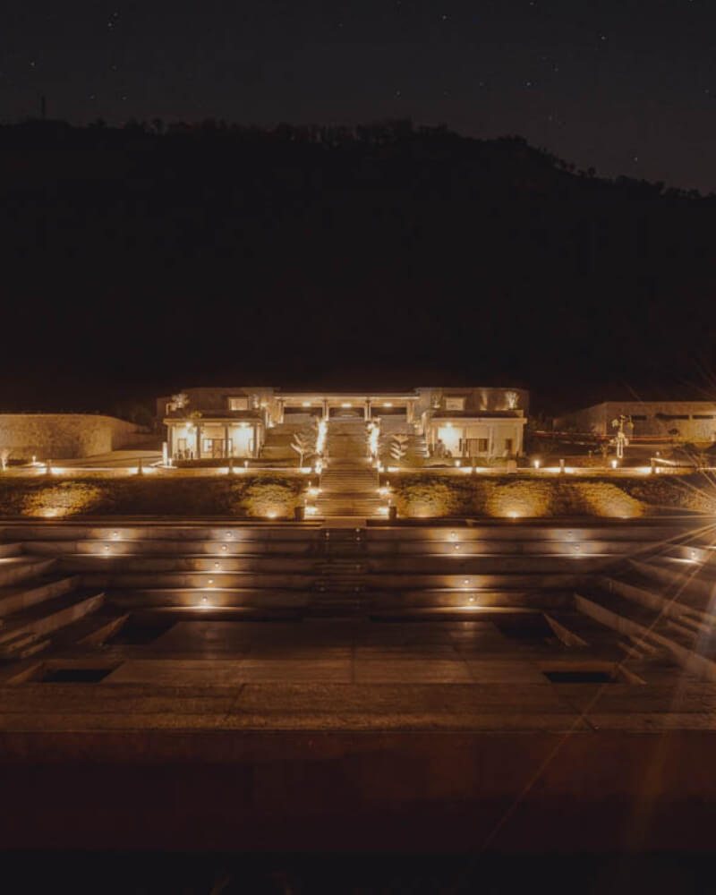 Amita Rasa at night from across the Kalyani — the amphitheatre steps picked out by lights set into the treads, a warm-lit stone building range with a central stair beyond, and the dark mass of the Nandi ridge against a star-flecked sky