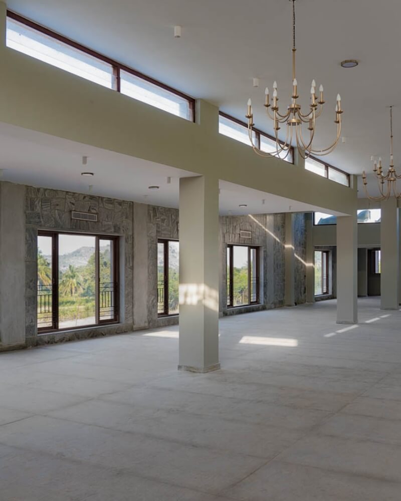 Inside the Aahara dining hall at Amita Rasa, unfurnished — a double-height room with a clerestory window band, brass candelabra chandeliers, rough stone-clad piers and full-height timber windows opening onto the hill and palms outside