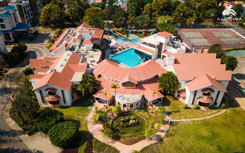 Aerial view of the main resort block at Aldovia (formerly Clarks Exotica) — red-tiled pitched roofs around a circular pavilion, a free-form blue pool with a yellow-and-blue waterslide behind it, tennis courts to the right, and a stone-and-water feature in a circular lawn at the front