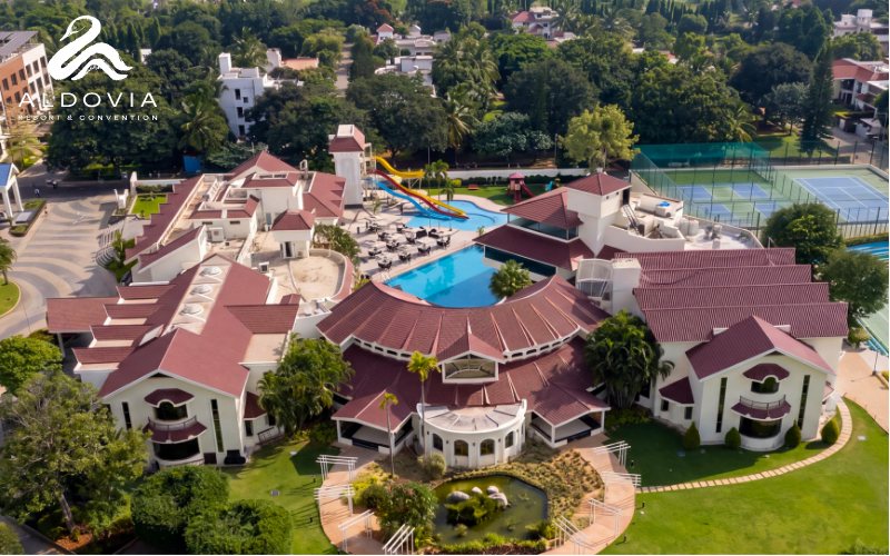 Higher aerial of Aldovia Resort & Convention showing the same wings under dark red roofs with the pool, waterslide, tennis courts and surrounding lawns, with neighbouring houses and trees beyond the boundary