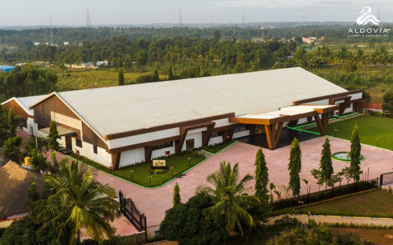 The standalone Ocean convention hall at Aldovia seen from the air — a very long single-span building with a pale roof and timber-clad angled entrance canopies, a brick-paved forecourt with a fountain and lamp posts, conifers along the boundary and open farmland behind