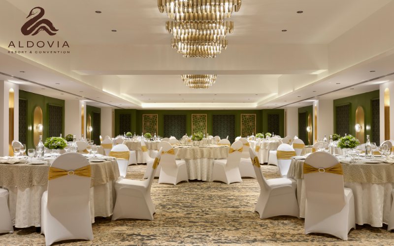 A ballroom at Aldovia set for a banquet — white-covered round tables with gold sashes and green hydrangea centrepieces under two tiered gold chandeliers, with sage-green fretwork wall panels and a cream-and-gold patterned carpet