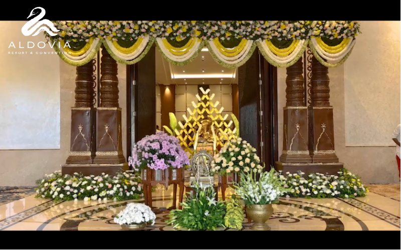 A wedding entrance set at Aldovia — carved dark timber pillars flanking open doors, hung with swagged garlands of yellow and cream flowers, with banks of white and pink blooms, purple chrysanthemums and brass urns on a polished inlaid marble floor and a gilded backdrop inside