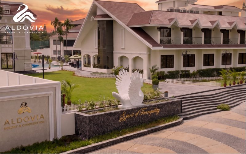 The entrance forecourt at Aldovia at sunset — a white swan sculpture on a black marble plinth lettered 'Spirit of Tranquility', a broad stepped terrace and paved drive, with the cream gabled resort block and a low white boundary wall carrying the Aldovia name behind