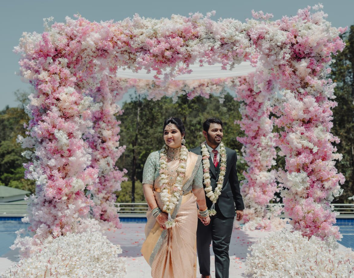 A couple standing under the blossom arch at the Coorg Marriott poolside after the ceremony, pool water visible either side
