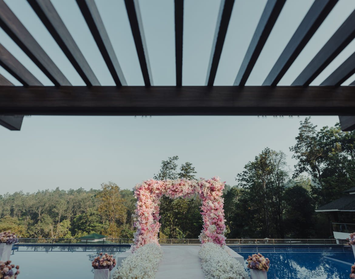 Pink and white floral arch standing on decking laid over the pool at Coorg Marriott, pergola beams overhead and the forest ridge beyond