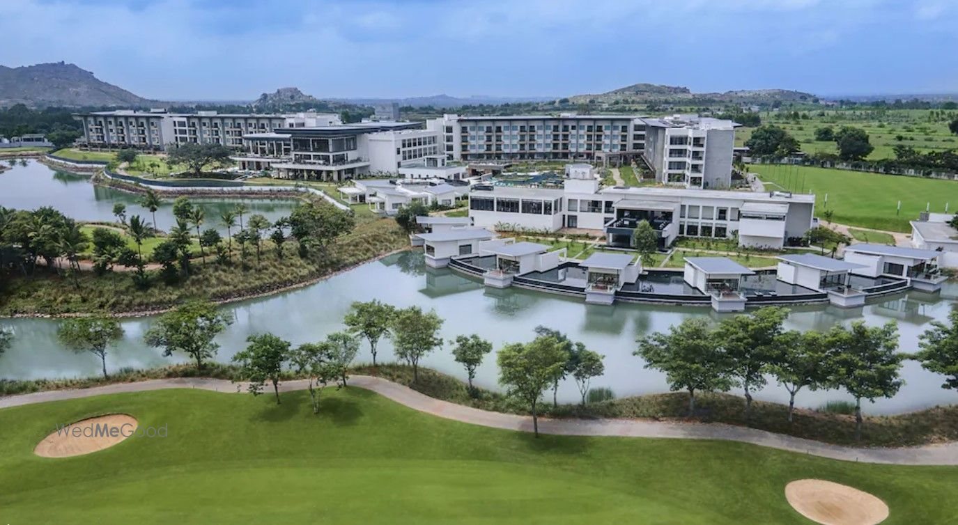 Aerial view of the whole resort — the low white room blocks and over-water pavilions wrapped around the lagoon, the green golf fairway and sand bunkers in the foreground and the Nandi Hills ridge on the horizon