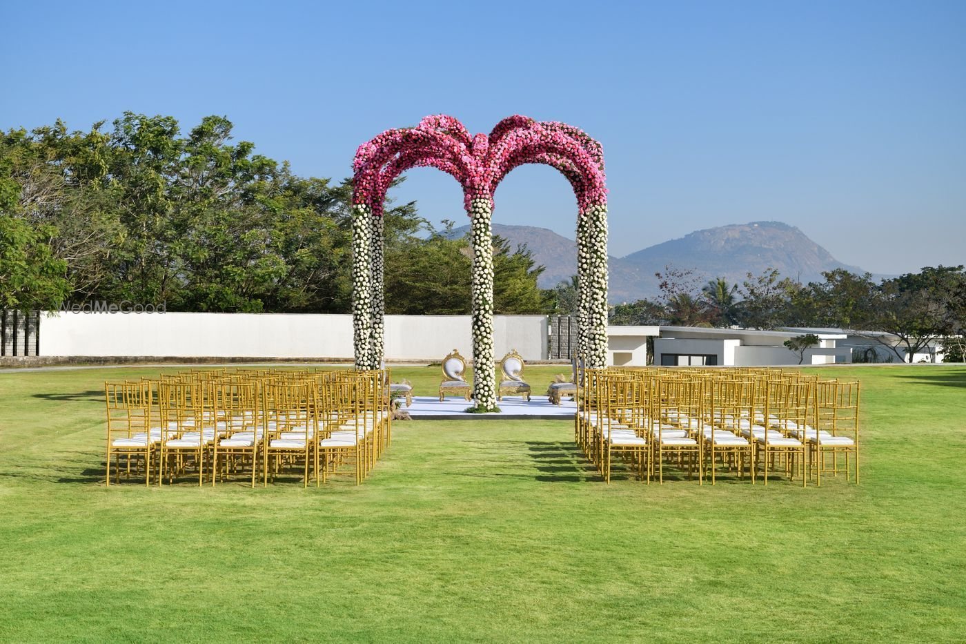 The Savannah Lawn set for a daytime ceremony — twin floral arches in pink and white roses over a white platform with two carved chairs, gold chiavari chairs ranked either side of a centre aisle, and the Nandi Hills rising behind the boundary wall