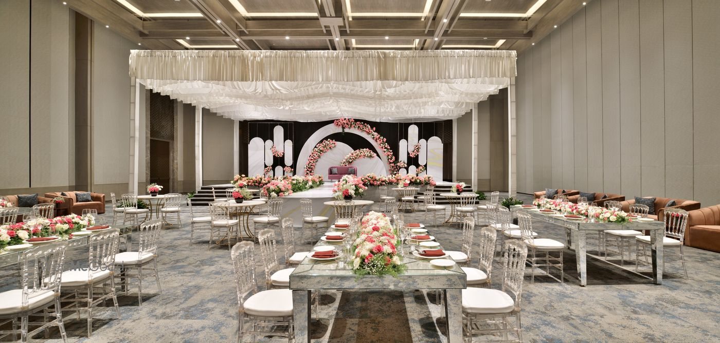 The Durbar hall dressed for a reception — a draped white ceiling canopy over the centre, an arched floral backdrop stage in blush and cream, mirrored long tables with ghost chiavari chairs, and the coffered ceiling grid and recessed lighting visible above