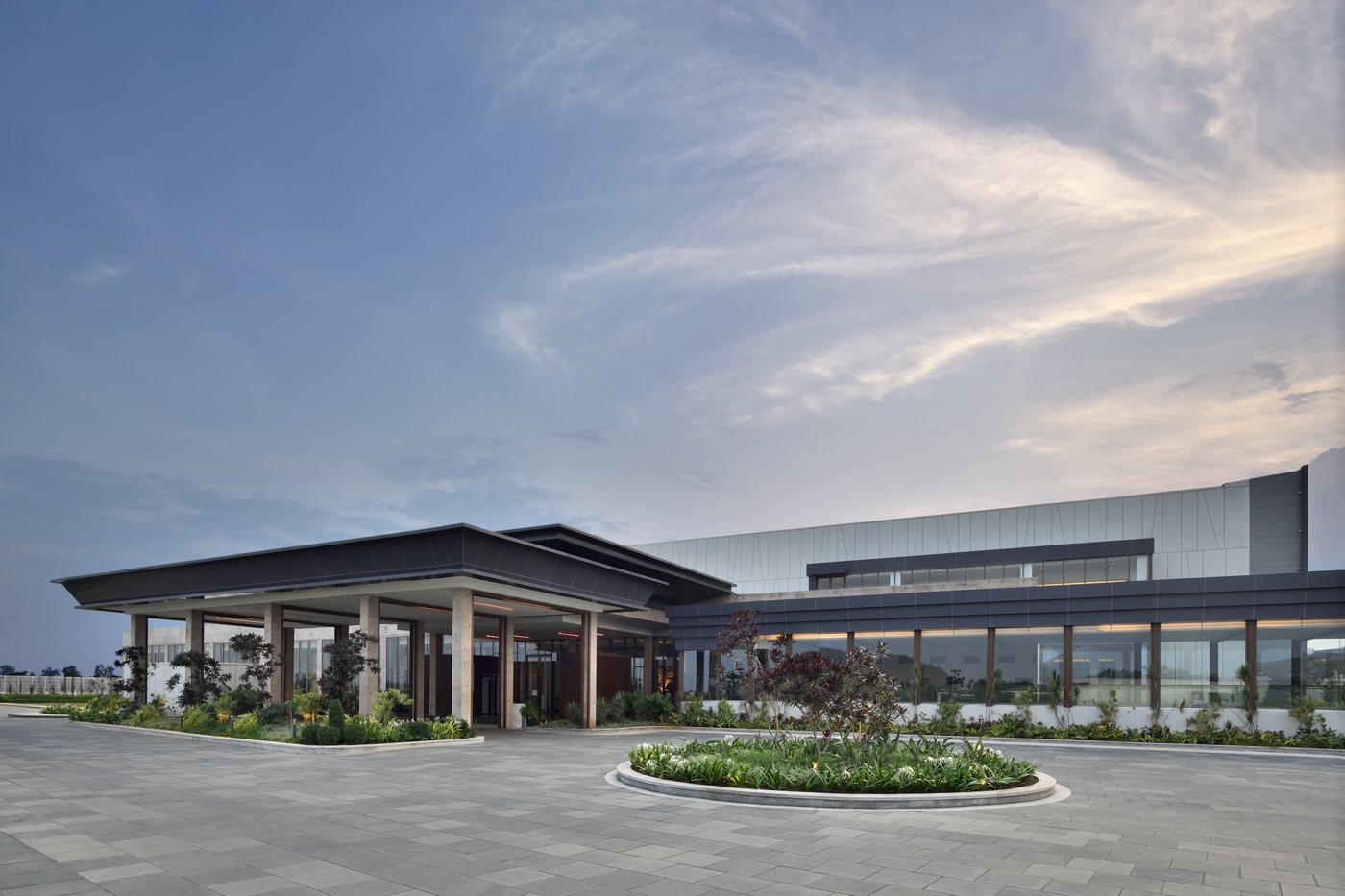 Exterior of the Durbar Convention Centre in early evening — a cantilevered porte-cochere over the entrance, a long glazed facade running away to the right and a planted circular island in the empty paved forecourt