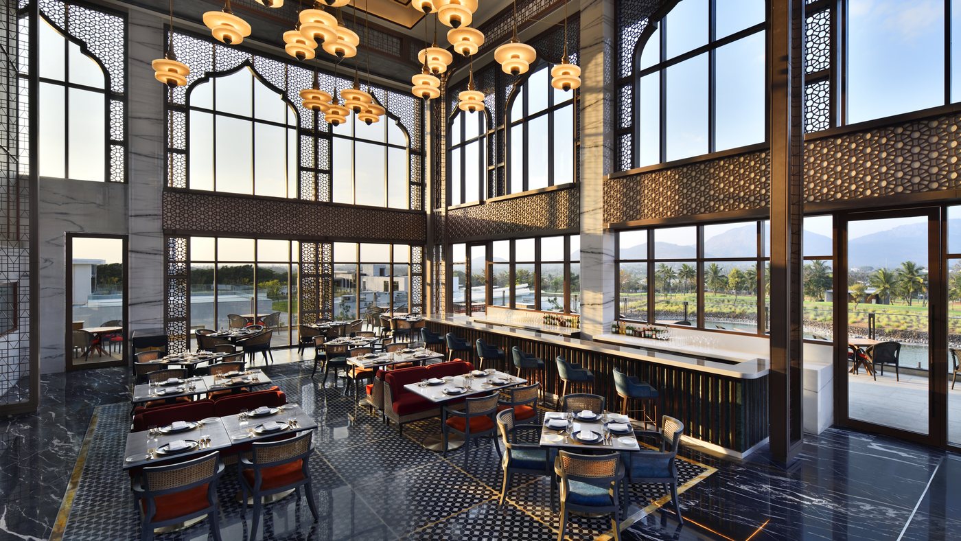 Interior of the Aaleeshan restaurant — a double-height dining room with Mughal-arched windows, pierced jaali screens, alabaster pendant chandeliers and black marble flooring, looking out to the fairway, lagoon and hills