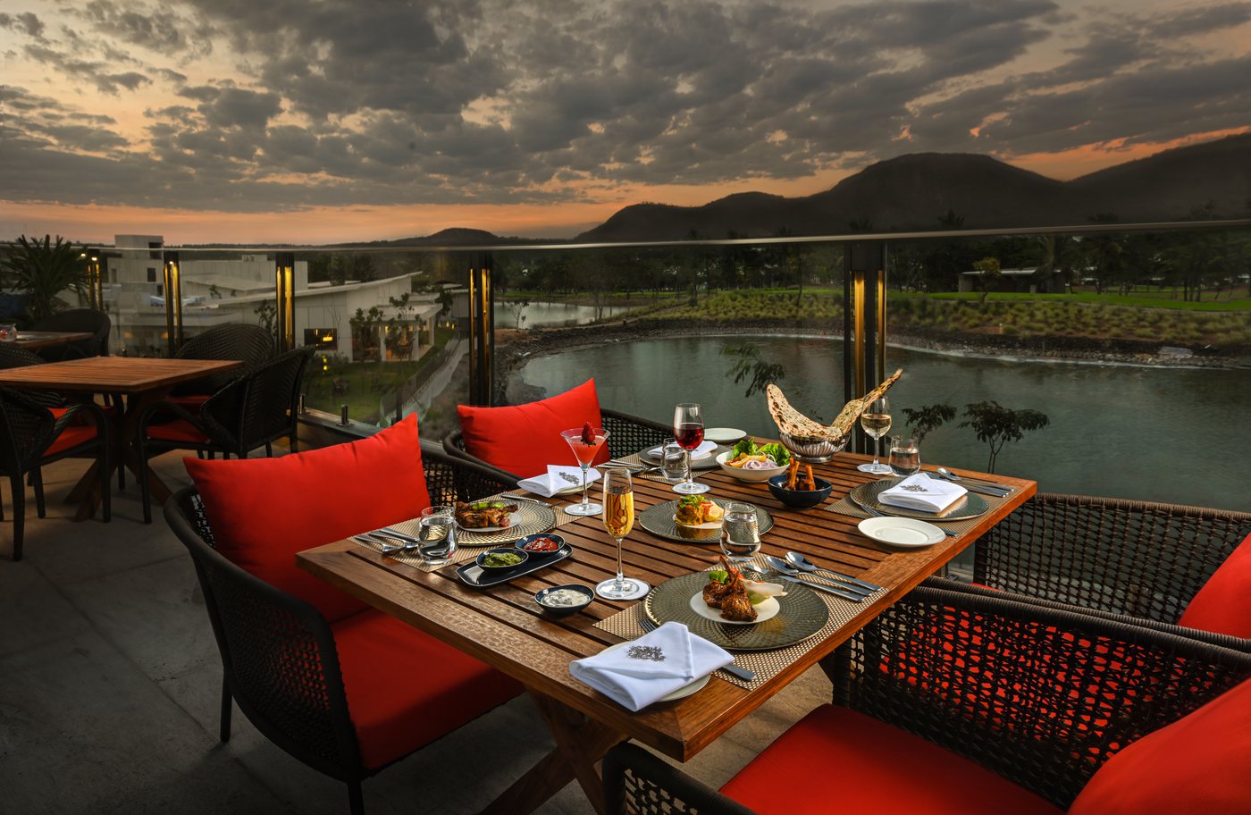 The Aaleeshan alfresco terrace laid for dinner at sunset — a timber table set with glassware and red cushioned wicker chairs, overlooking the lagoon and fairways with the Nandi Hills silhouetted under an orange and grey sky