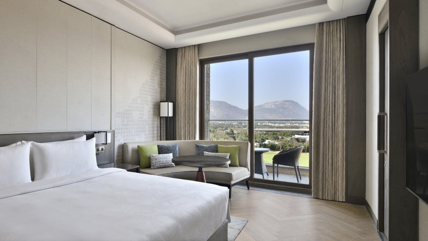 A corner guest suite with a king bed, a curved banquette and a full-height sliding window onto a private balcony, looking straight out at the Nandi Hills massif with the green golf fairway below