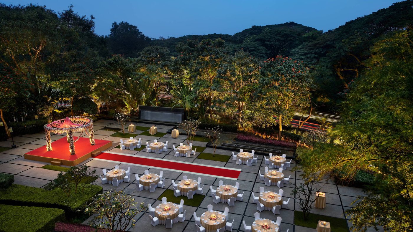 The JW Lawns at JW Marriott Bengaluru set for a wedding at dusk — a floral mandap on a raised red platform, a red carpet aisle and round dining tables laid across a checkerboard terrace of stone slabs and turf, with the dense Cubbon Park tree canopy behind