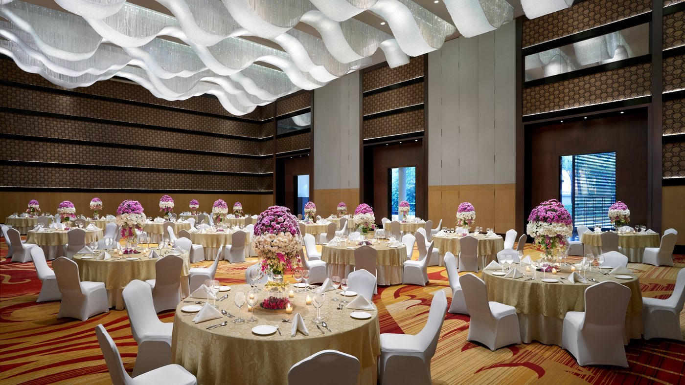 The Grand Ballroom at JW Marriott Bengaluru laid with banquet rounds in gold and white, tall pink and white floral centrepieces, and a sculptural white wave-form ceiling installation over crystal strands