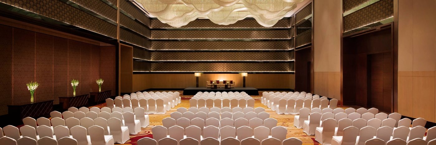 The Grand Ballroom at JW Marriott Bengaluru set theatre-style with rows of white-covered chairs facing a low stage, under the double-height panelled walls and the wave-form ceiling