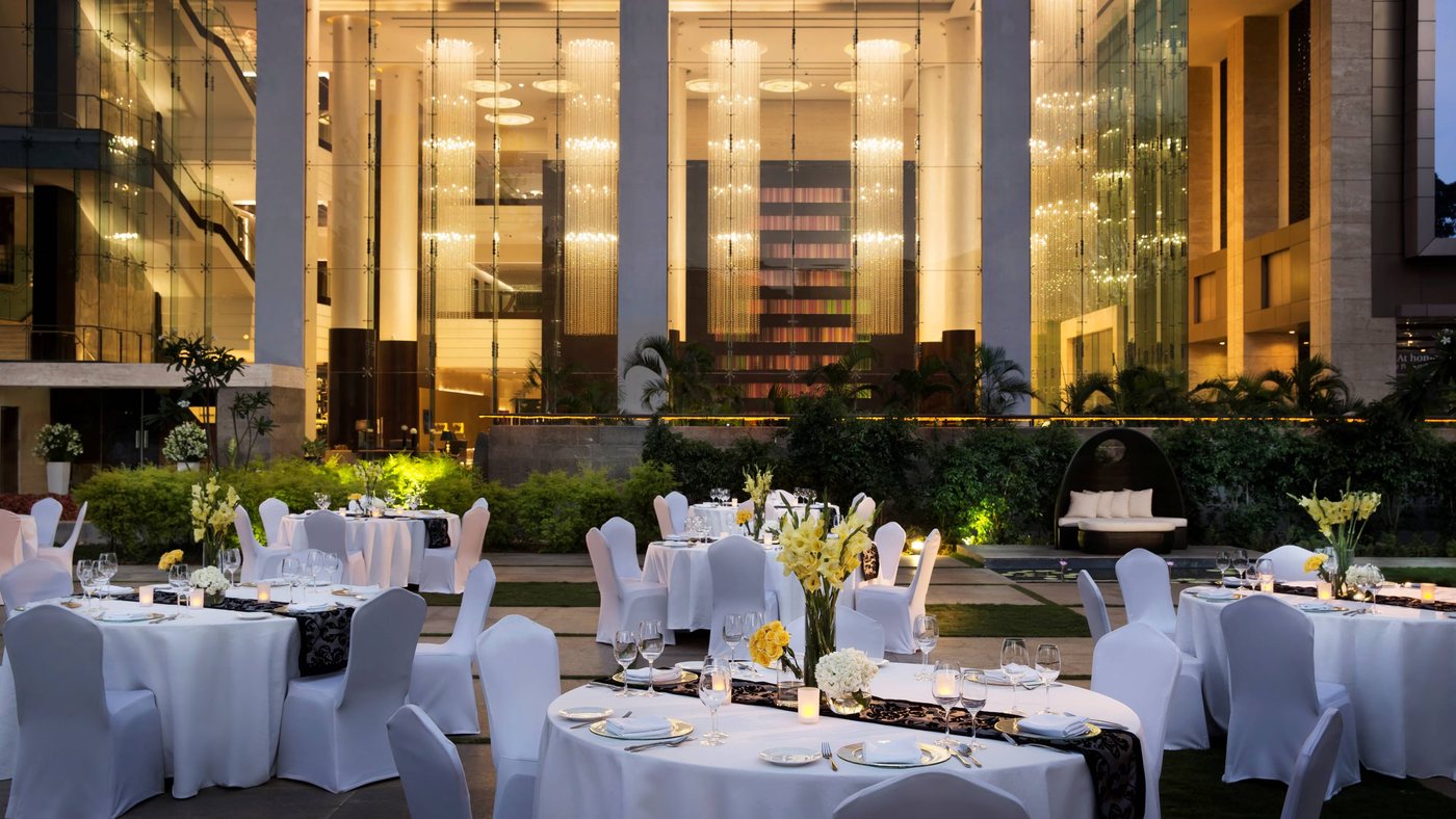 An outdoor dinner laid in the courtyard at JW Marriott Bengaluru — round tables in white with yellow gladioli, set against the hotel's glazed atrium and its full-height cascading crystal-strand chandeliers